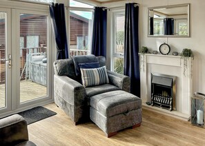A living room with a grey sofa, a fireplace, a clock, and a view of the outdoors. - Country Holiday Home Four (Tintagel)