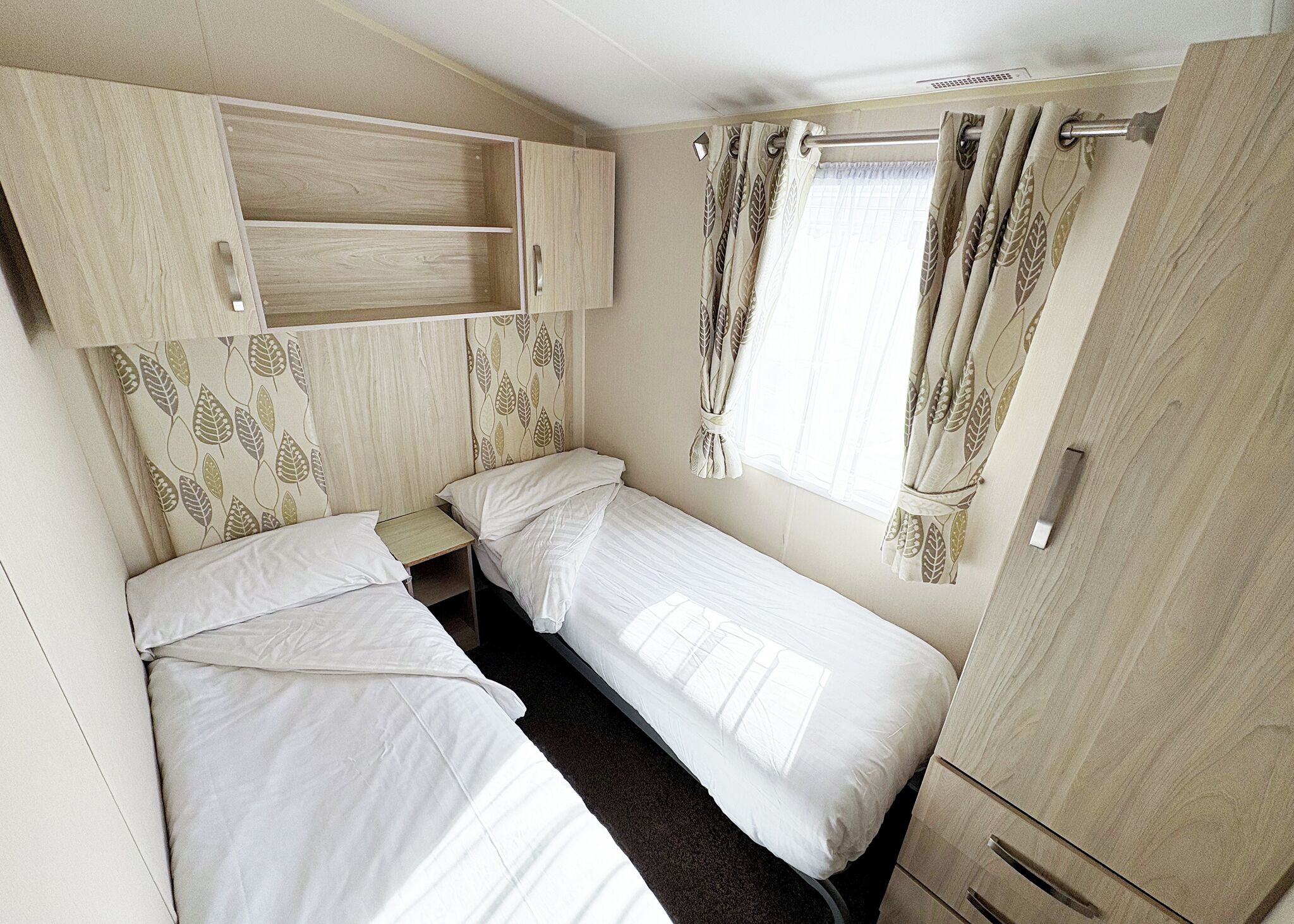 Una habitación compacta con dos camas individuales, una mesita de noche pequeña, un armario y una ventana con cortinas.