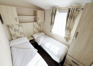Une chambre de taille modeste, équipée de deux lits simples, d’une petite table de chevet, d’une armoire et d’une fenêtre avec des rideaux.