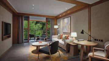 Una habitación de hotel moderna con sofá, dos sillas, una mesita, televisor y balcón con cortinas.