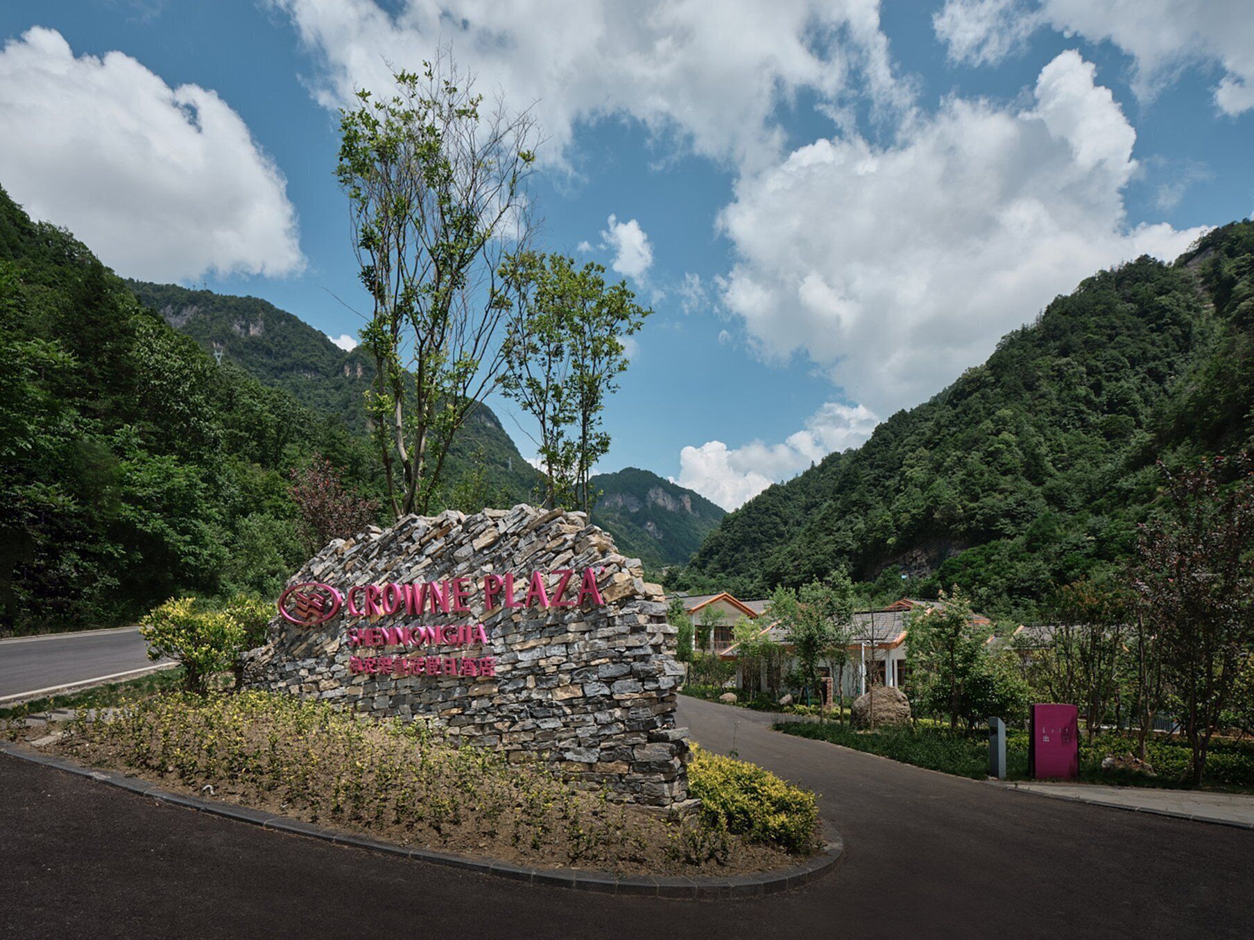 Un letrero de piedra con la inscripción "CROWNE PLAZA" y "SHENZHEN", rodeado de follaje y montañas.