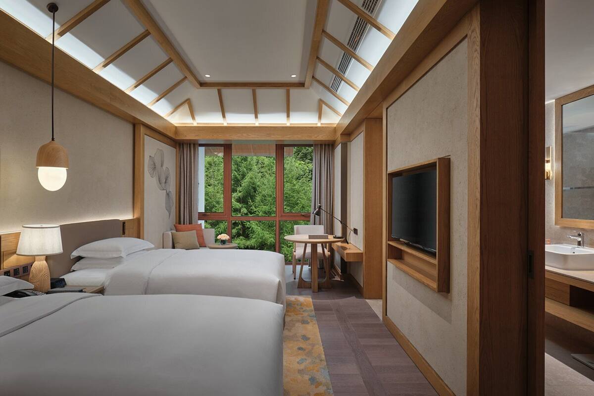 Habitación de hotel con dos camas, un escritorio, televisor y un amplio ventanal con vistas a zonas verdes.