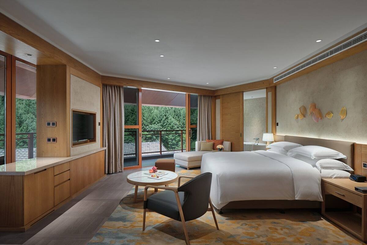 Habitación de hotel moderna con una cama grande, un escritorio, una silla y un balcón con vistas a zonas verdes.
