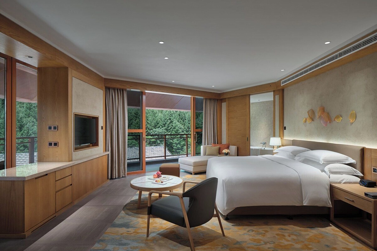 Une chambre d’hôtel moderne dotée d’un grand lit, d’un bureau, d’une chaise et d’un balcon offrant une vue sur la verdure.