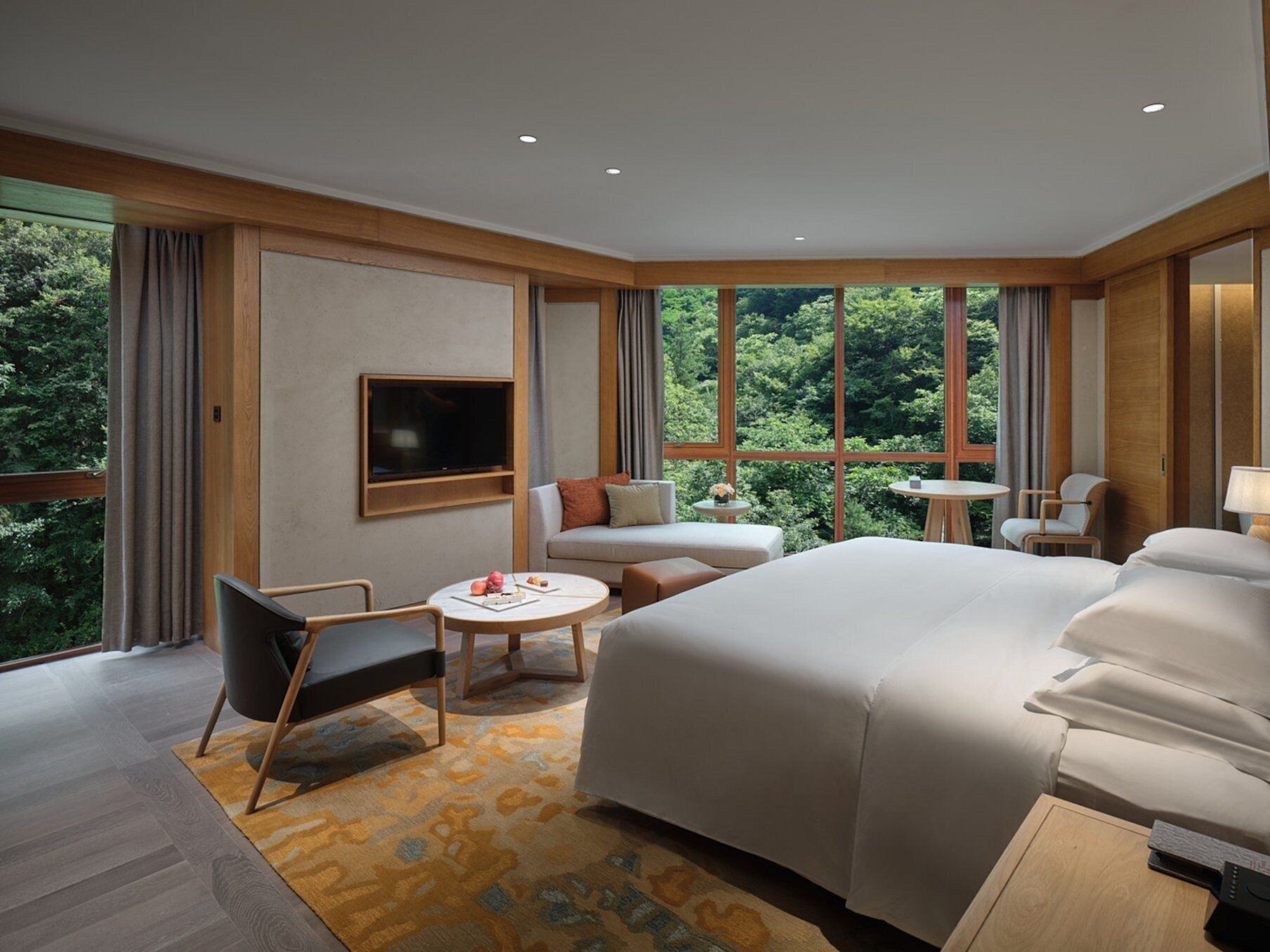 Una habitación de hotel moderna con una cama grande, un sofá, un televisor montado en la pared y vistas al bosque a través de amplias ventanas.