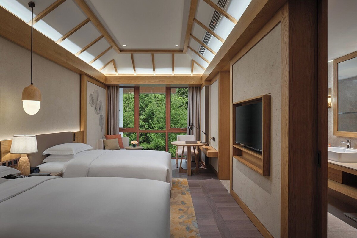 Une chambre d’hôtel avec deux lits, un bureau, une télévision et une grande fenêtre donnant sur un paysage verdoyant.