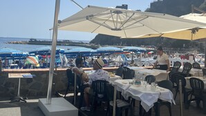Un área de comedor al aire libre con mesas y sillas, sombrillas y vistas al mar y a las embarcaciones.