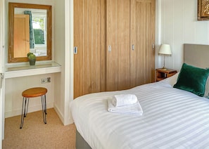 A bedroom with a bed, a nightstand, a lamp, a mirror, a wooden wardrobe, and a small stool.