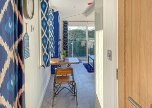 Un couloir décoré de rideaux à motifs, une chaise en bois, une table ornée d’un pot de plantes et une porte-fenêtre coulissante donnant sur un espace extérieur.