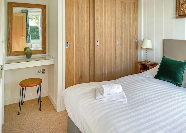 Quarto com cama, mesa de cabeceira, luminária, espelho, guarda-roupa de madeira e um pequeno banquinho.