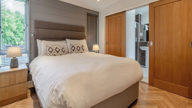 Un dormitorio con una cama grande, lámparas de noche, un armario de madera y un baño visible a través de una puerta de cristal.