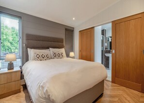A bedroom with a large bed, bedside lamps, a wooden wardrobe, and a bathroom visible through a glass door.
