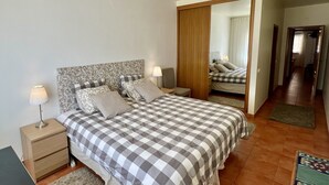 A bedroom with a checkered bedspread, a nightstand with a lamp, a wooden wardrobe, and a mirror reflecting the room. - Carvoeiro Terrace - Gorgeous apartment close to beach (Carvoeiro)