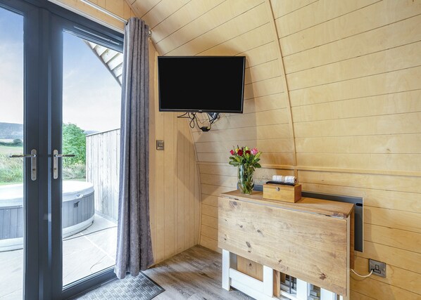 Interior em madeira com porta de vidro deslizante, televisão montada, cómoda de madeira e um vaso com flores.