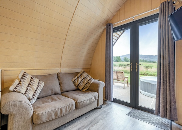 A cozy room with a wooden sloped ceiling, a sofa with patterned cushions, and a sliding glass door leading to an outdoor seating area. - 1 bedroom accommodation in Great Busby (Great Busby)