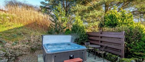 Un jacuzzi con un banco de madera y una pequeña mesa, rodeado de follaje y un muro de piedra.