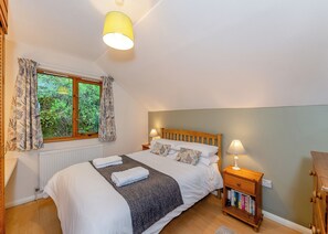 Un dormitorio con cama, ventana, radiador, mesita de noche con lámpara y una mesita auxiliar de madera con libros.