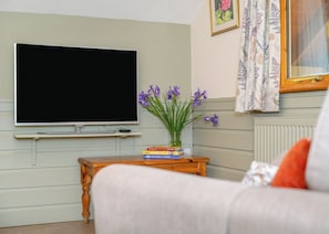 Uma sala de estar com televisão de tela plana, uma mesinha de madeira com livros, um vaso com flores e um sofá.