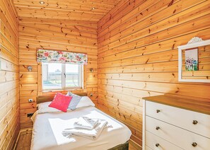 Une pièce aux murs lambrissés, comprenant un lit, une commode, une fenêtre ornée d’un rideau à motifs floraux et offrant une vue sur l’extérieur.
