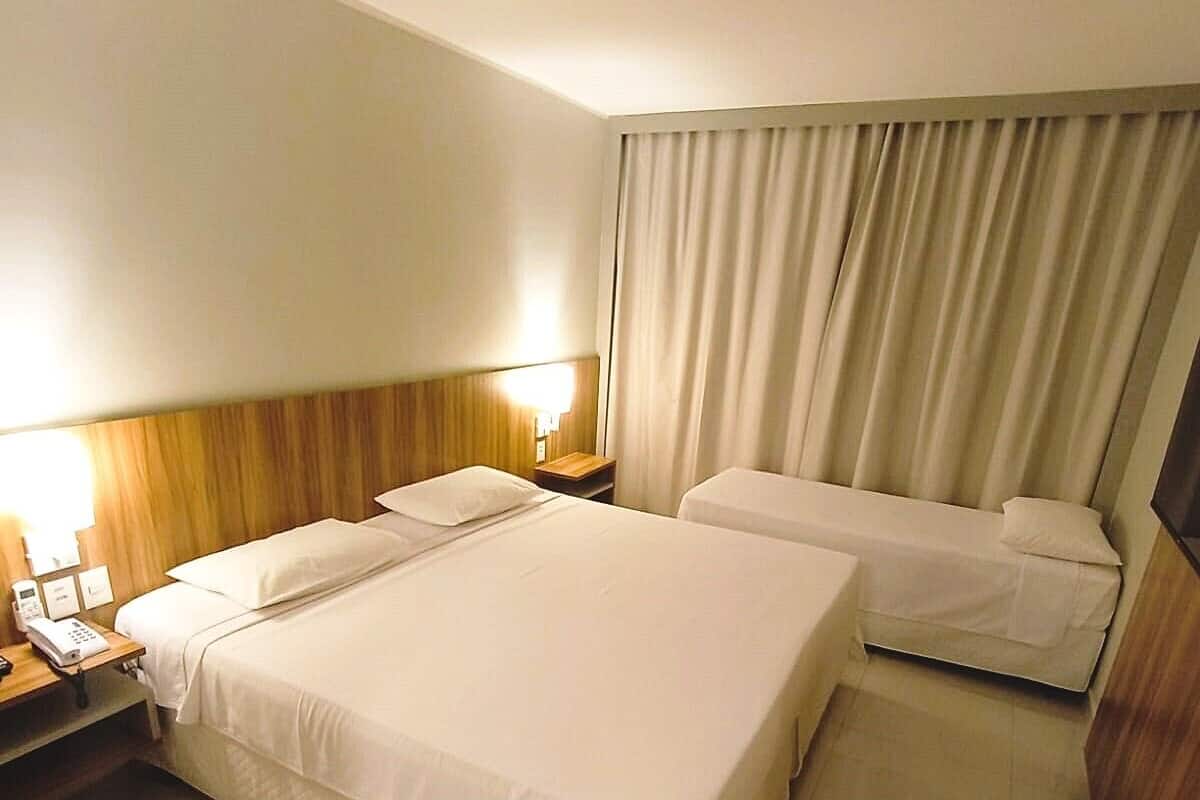 A hotel room with two beds, a wooden headboard, a bedside table with a lamp, and a curtain.