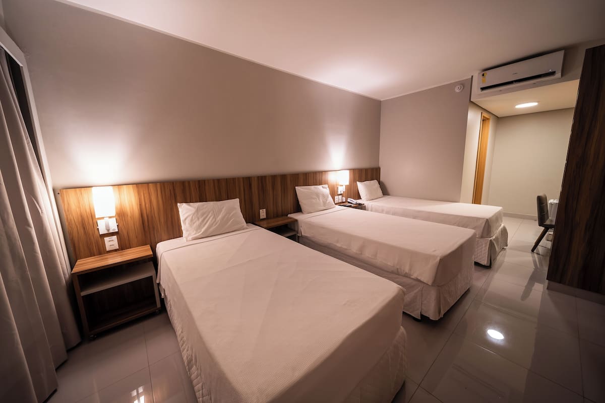 A hotel room with three single beds, a wooden headboard, a nightstand, a desk, and an air conditioning unit.
