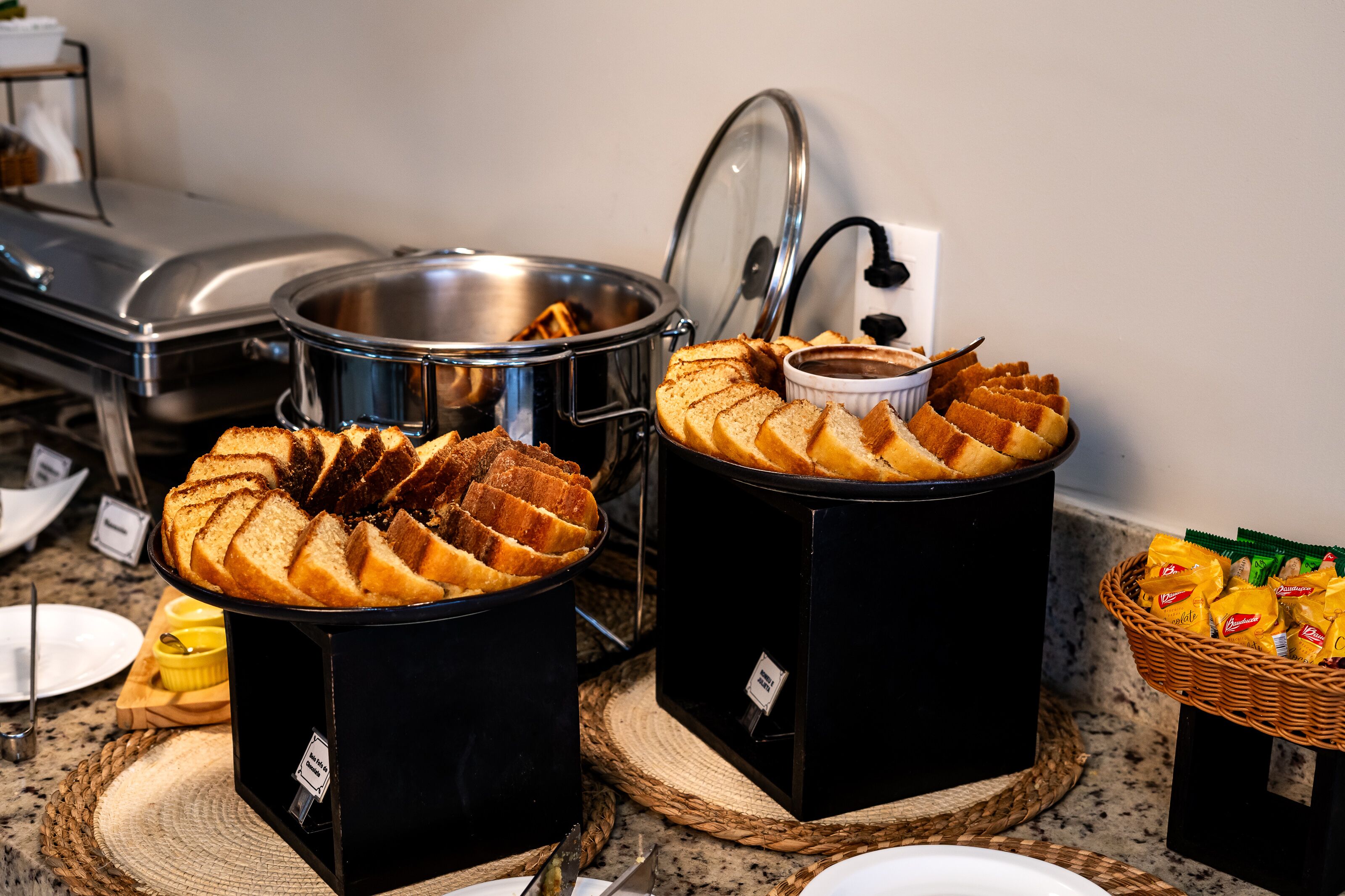 Disposição de buffet com diversos alimentos, incluindo pães e doces, dispostos sobre um balcão.
