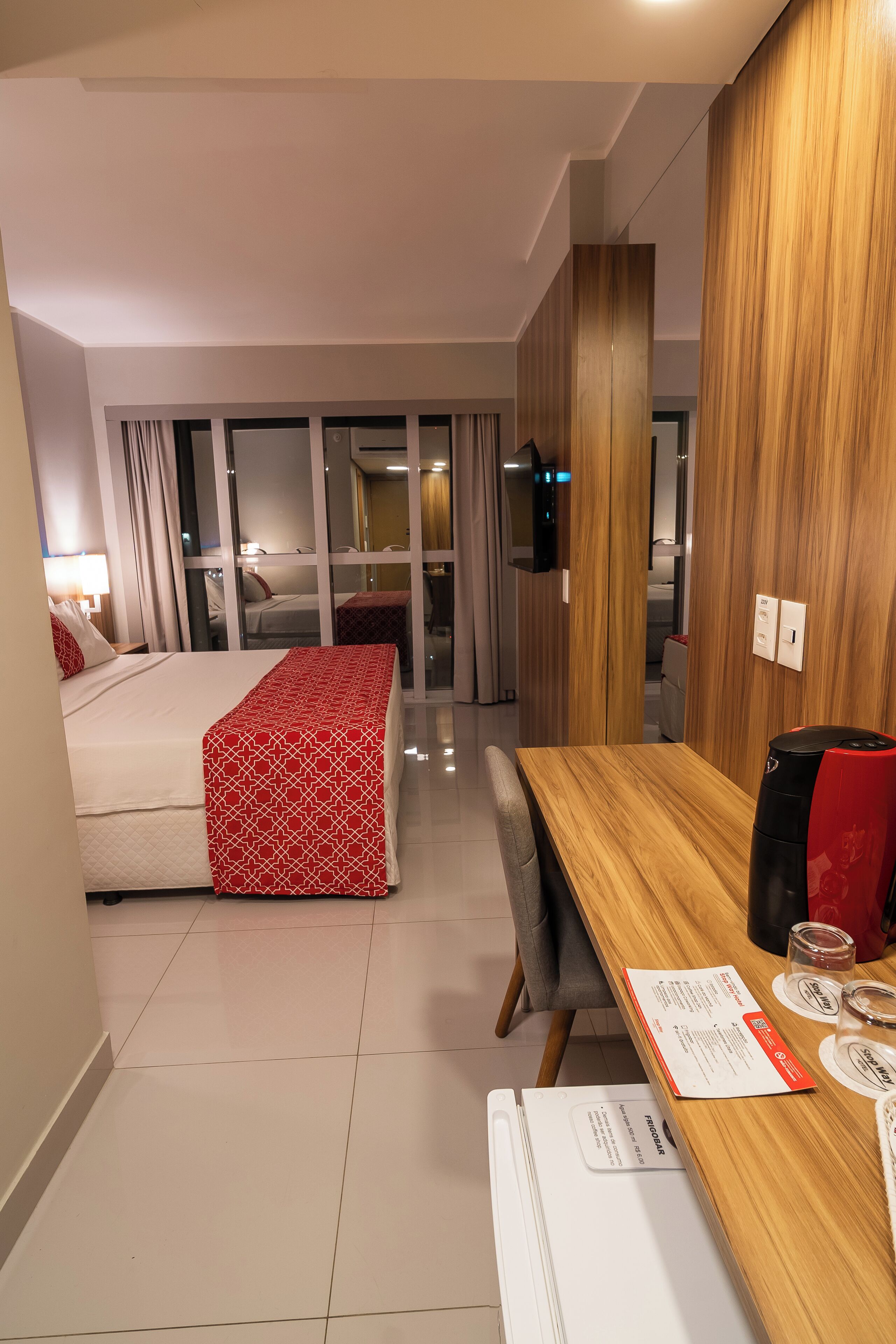 A hotel room with a bed, a wooden table, a desk, a chair, a TV, and a sliding glass door leading to a balcony.