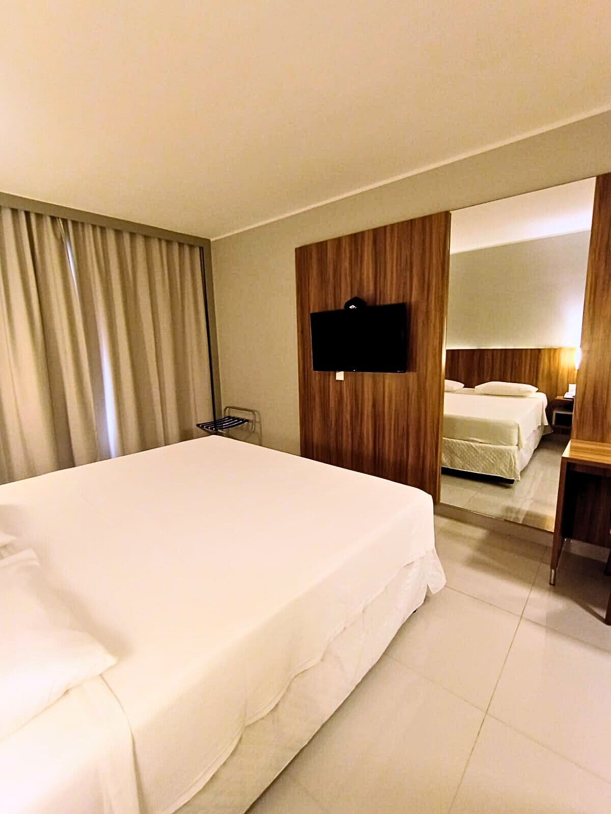 Ein Hotelzimmer mit einem großen Bett, einem an einer Holzvertäfelung befestigten Fernseher und einem Spiegel, der den Raum reflektiert.