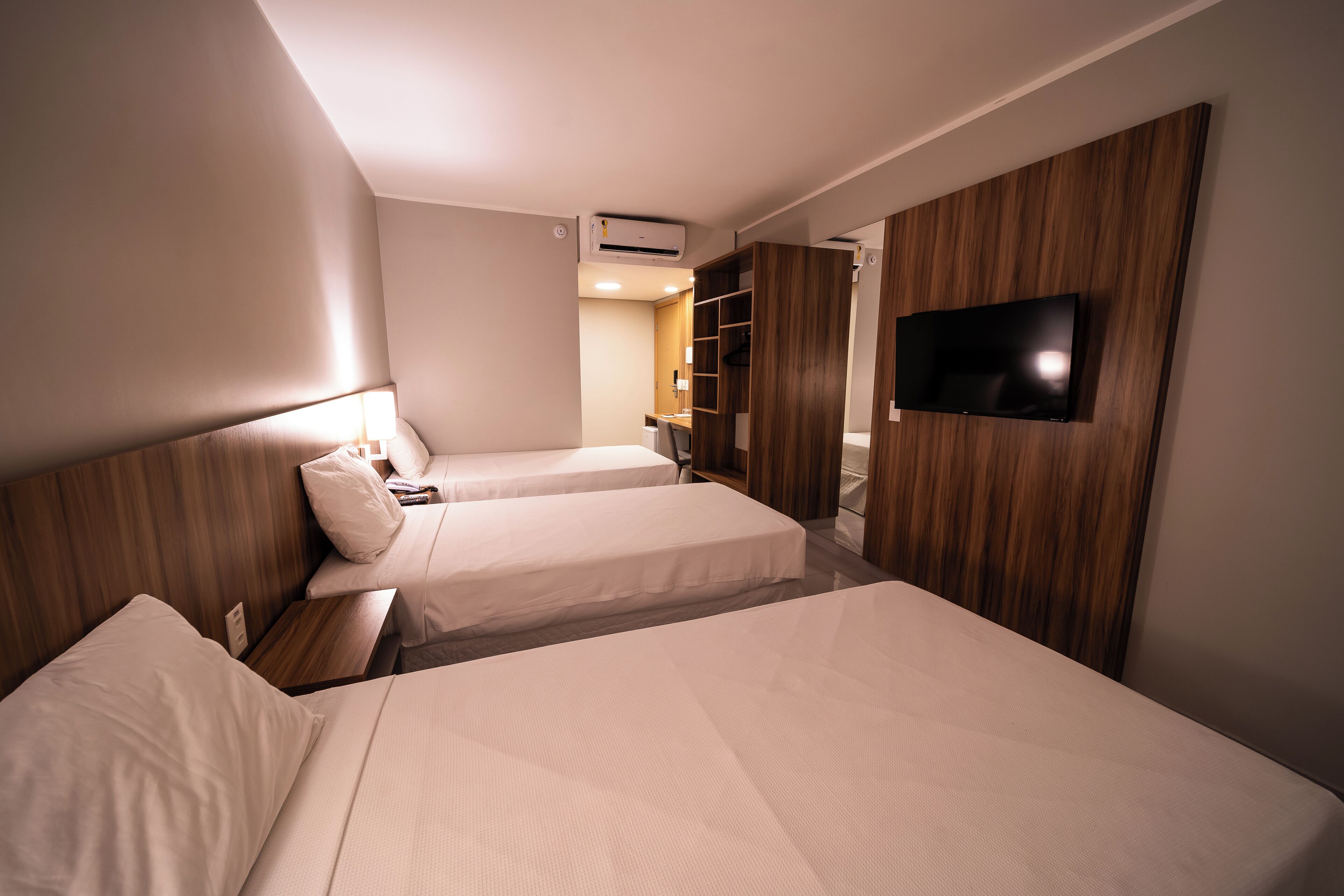 Habitación de hotel con dos camas, un televisor montado en la pared, un armario de madera y una mesita de noche con lámpara.
