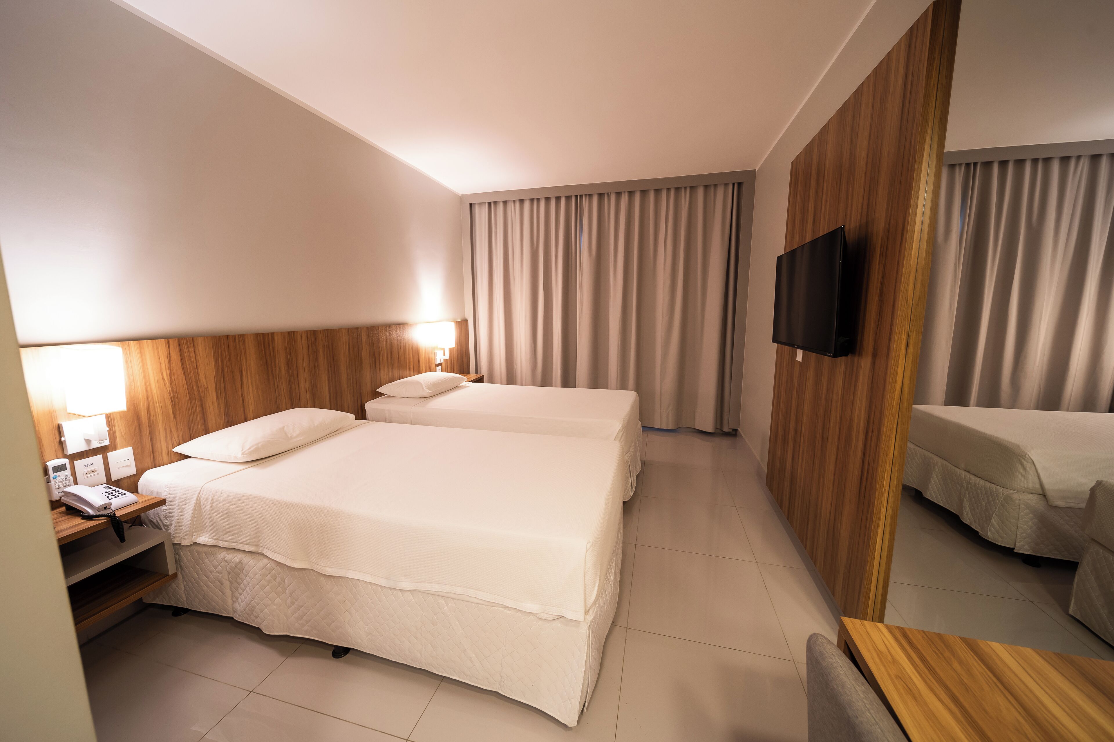 Een hotelkamer met twee bedden, een aan de muur bevestigde televisie, een nachtkastje met een telefoon en een houten paneel aan de muur.