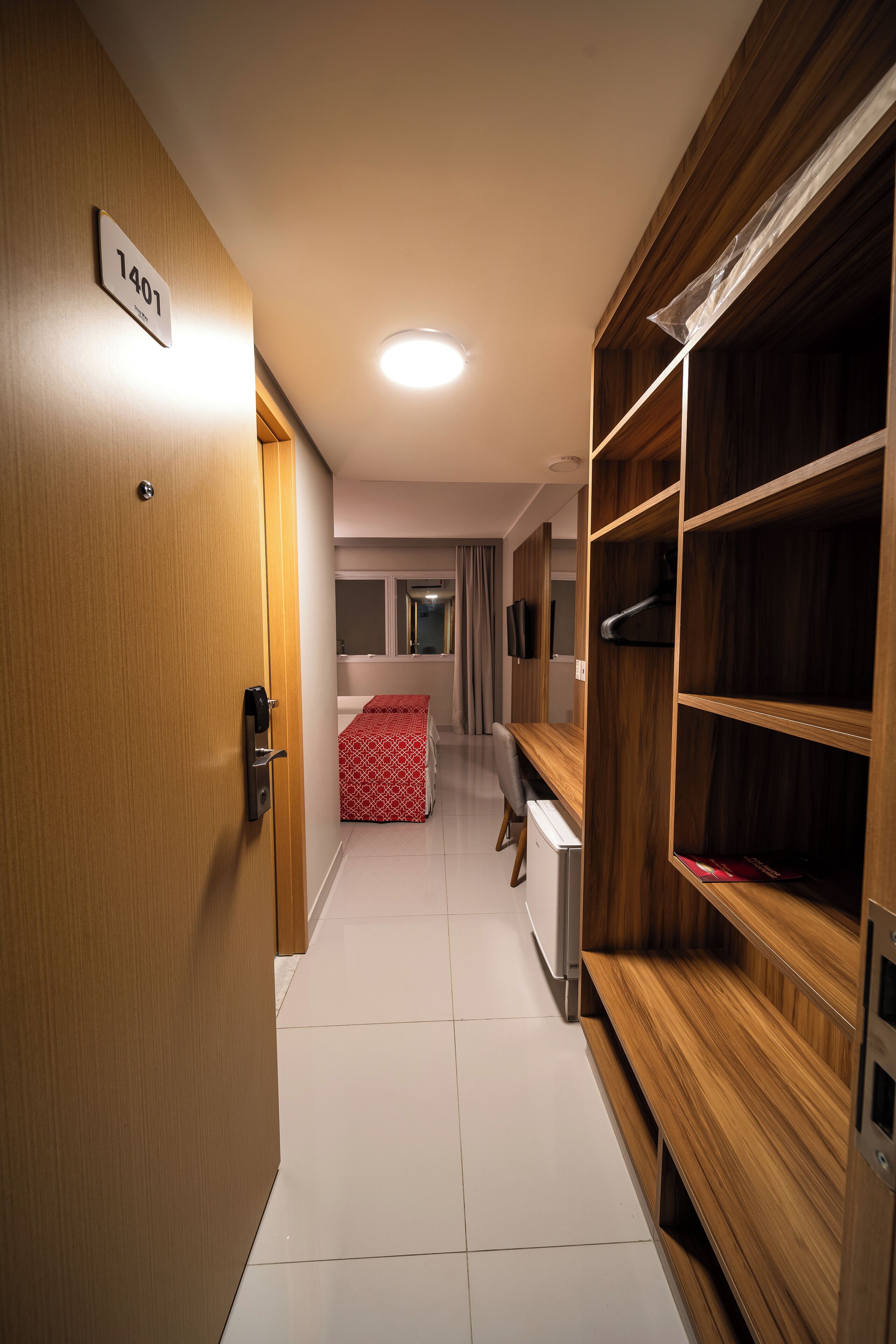 A hotel corridor with a wooden door labeled "1407", a bench, and a bed with a red and white patterned bedspread.