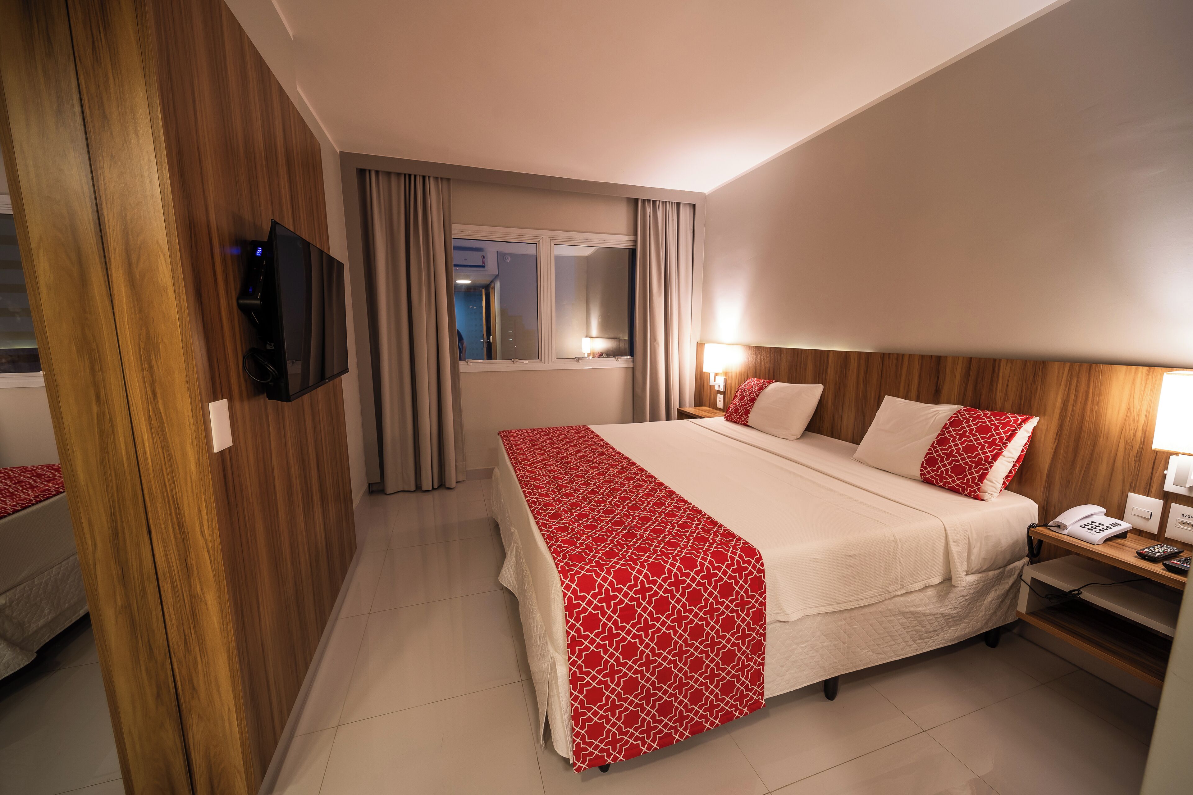A hotel room with a large bed, a TV mounted on the wall, a bedside table with a lamp, and a window with curtains.