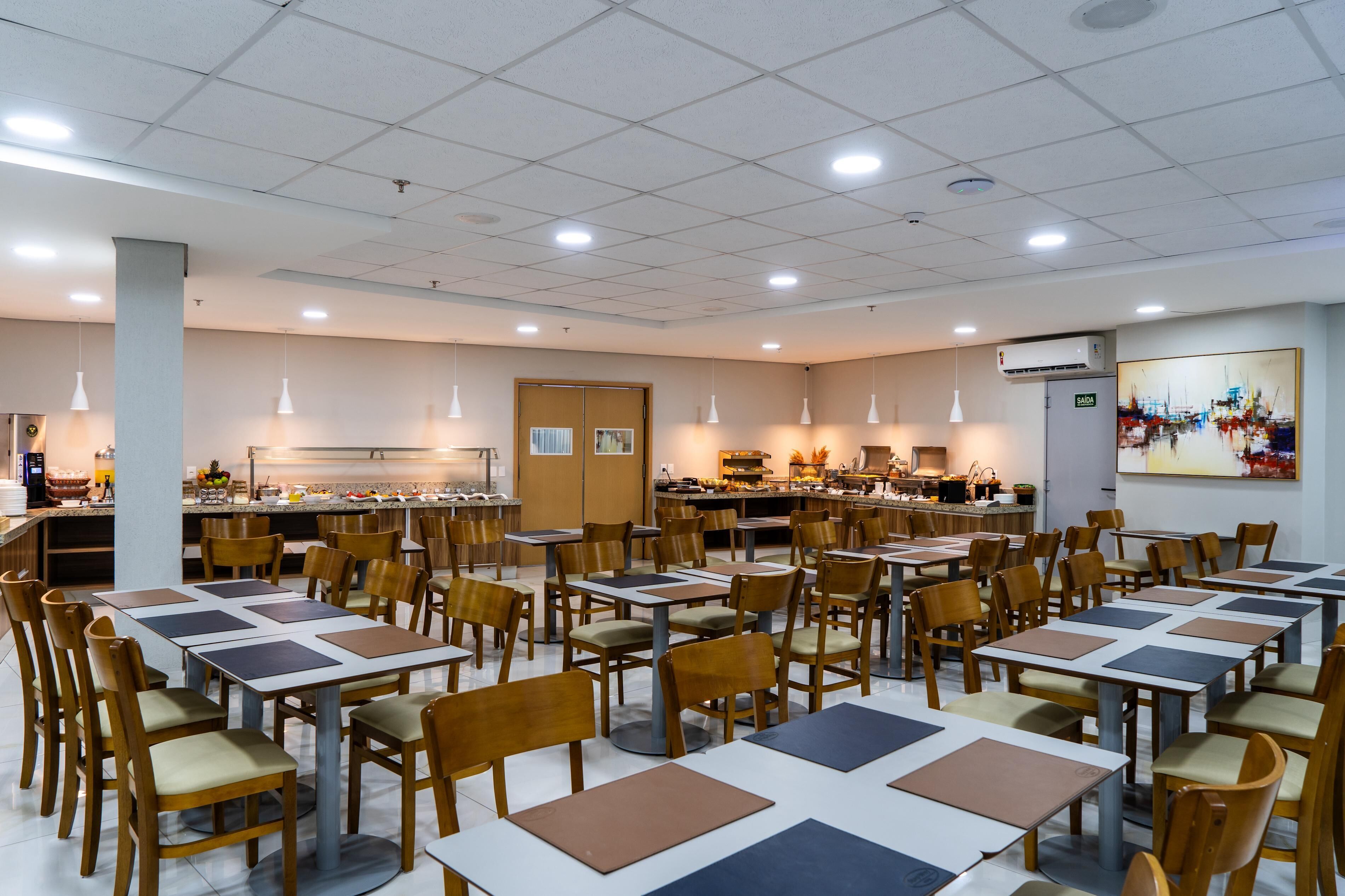 A spacious dining area with multiple tables and chairs, a kitchen in the background, and a painting on the wall.