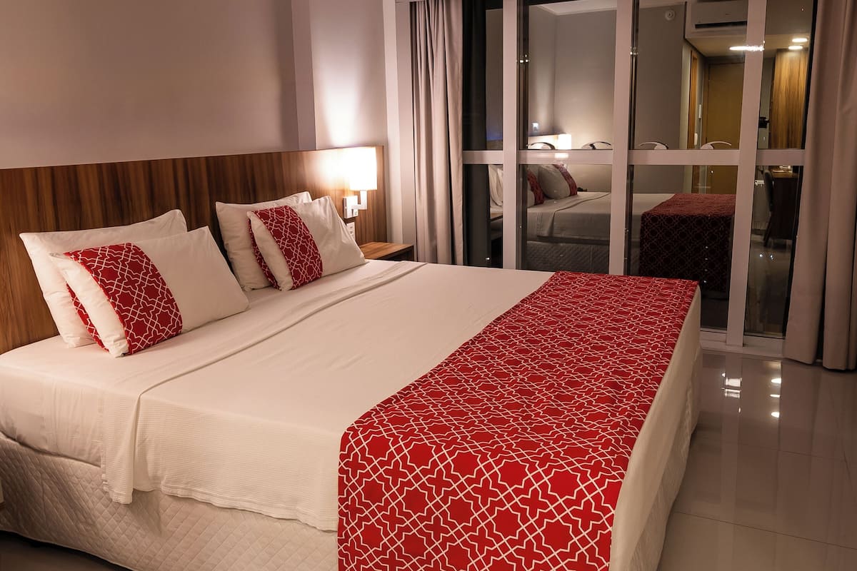 A hotel room with a large bed, a nightstand, a lamp, and a sliding glass door leading to a balcony.