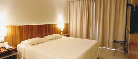 A hotel room with a large bed, a desk with a lamp, a chair, and a TV mounted on the wall.