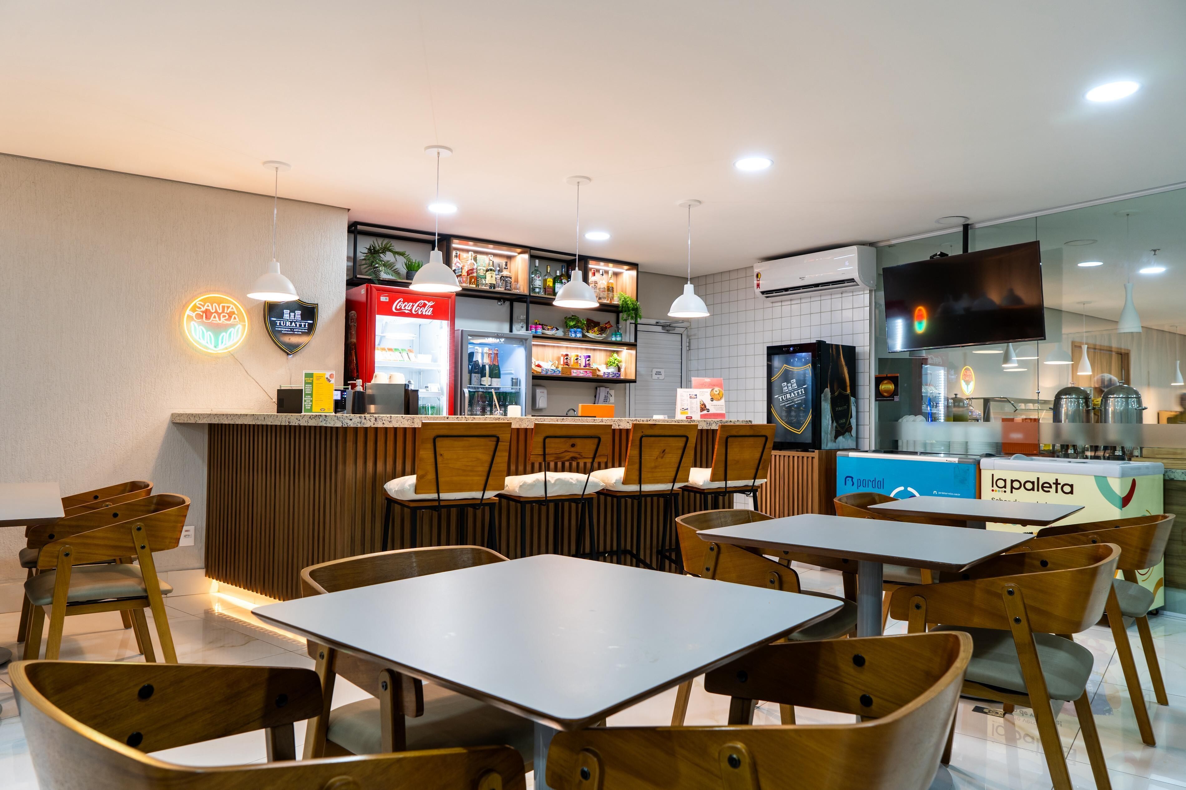 A restaurant interior with wooden chairs and tables, a counter with a Coca-Cola refrigerator, and a television mounted on the wall.