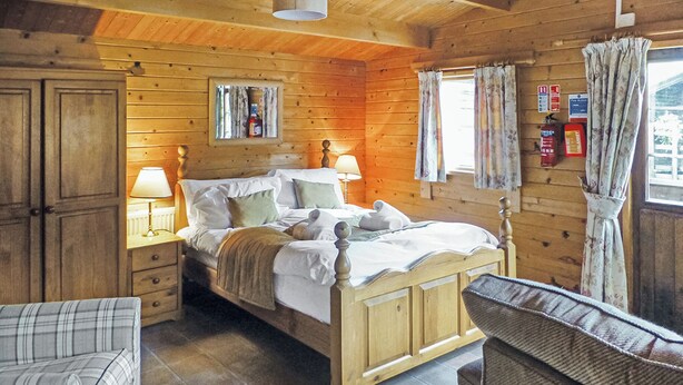 A wooden cabin bedroom with a bed, bedside tables, a chair, and a window with curtains. - Studio timber lodge with one double. (Kentisbeare, Nr Cullompton)