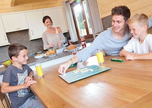 Eine Familie isst an einem Holztisch und spielt ein Brettspiel, dazu steht ein Glas Orangensaft und eine Wasserflasche.