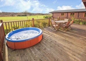 A hot tub on a wooden deck with a table and chairs, surrounded by a wooden fence and a view of a green field.