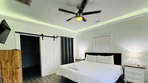 Ein Schlafzimmer mit Bett, Nachttisch, an der Wand montiertem Fernseher, Deckenventilator und Holzschrank.