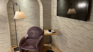 Una stanza con una poltrona viola, una lampada da terra, un tavolino con un telecomando e una TV fissata al muro.