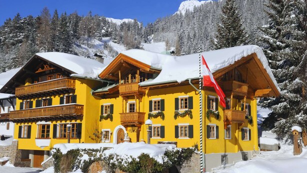 Un edificio giallo con balconi e una bandiera rossa, circondato da alberi e montagne innevate.