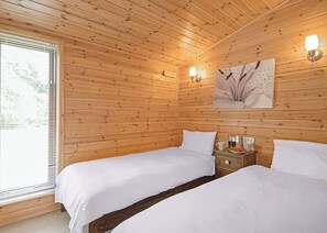 Quarto com paredes de madeira, duas camas, uma mesa de cabeceira com abajur e uma janela com persianas.