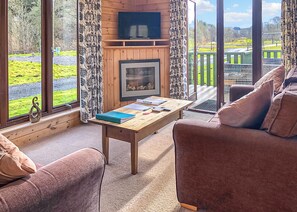 A cozy room with a fireplace, a sofa, a table with books, and a view of a green landscape.