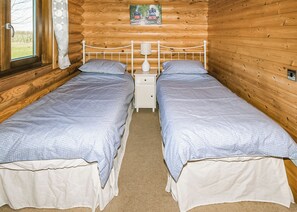A wooden cabin room with two single beds, a nightstand with a lamp, and a window with curtains.