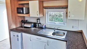 A compact kitchenette with a built-in microwave, toaster, and coffee maker, a small refrigerator, a sink with a window above it, and wooden cabinets. - The Gold Plus 2 caravan (pet) is 12ft wide with one double and one twin. (Clarach Bay, Nr Aberystwyth)