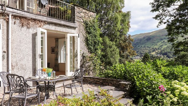A stone house with a balcony, a table with chairs, and a garden with various plants and flowers.