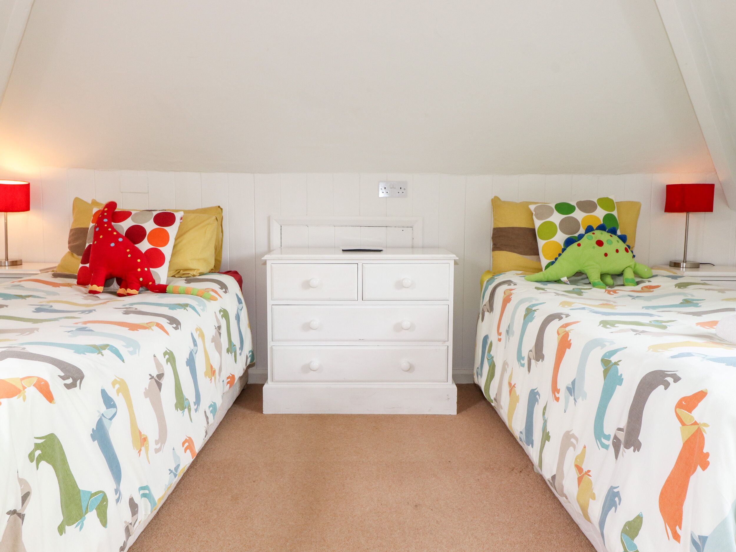 A bedroom with two beds, each with a dinosaur-themed bedspread, a white dresser, and red lamps on either side.