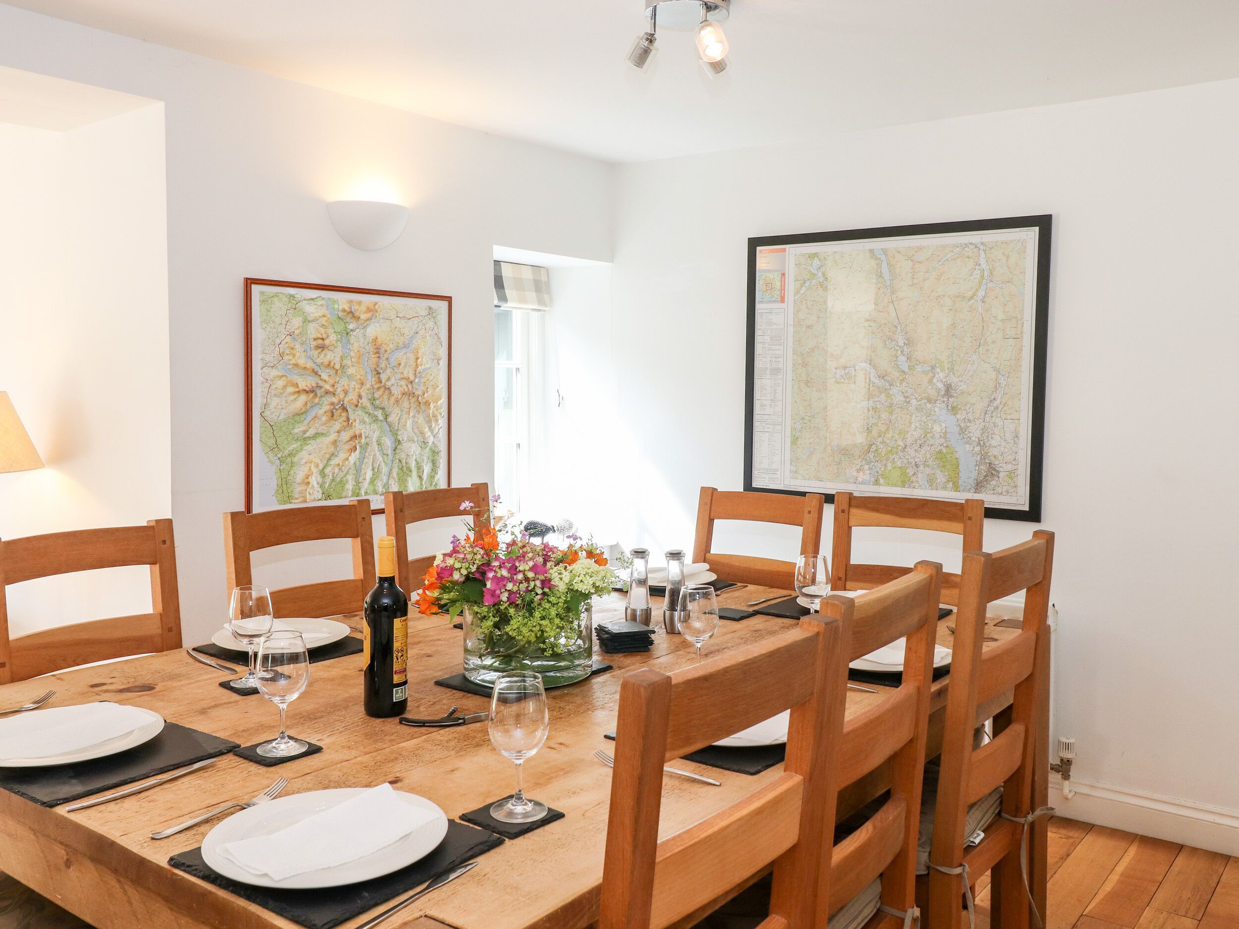 A dining area with a wooden table set for six, surrounded by wooden chairs. There are two framed maps on the walls, a bottle of wine, and a vase with flowers on the table.