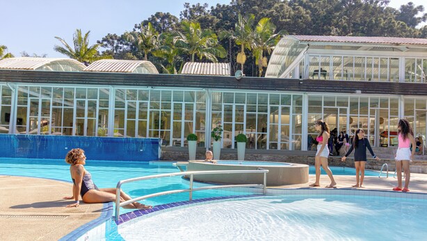 Una piscina con agua azul cristalina, rodeada por un recinto de cristal con palmeras y un edificio con techo metálico.